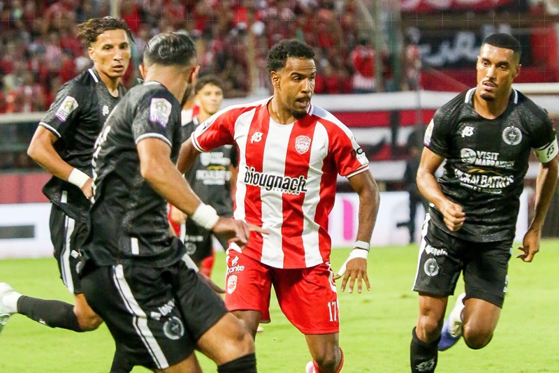 لاعب كرة قدم يرتدي قميصا مخططا باللونين الأحمر والأبيض من فريق الكوكب المراكشي يركض بالكرة في الملعب.